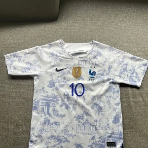 Frankrike Mbappé #10 Nike fotbollströja - Snygg Frankrike fotbollströja från Nike med Mbappé och nummer 10 på ryggen. Tröjan är vit med ljusblå mönster, har korta ärmar och är tillverkad i lätt, ventilerande material. FIFA-märke och FFF-logga på bröstet. Perfekt för dig som älskar landslagsfotboll! Tröjan är för ca 12-13
