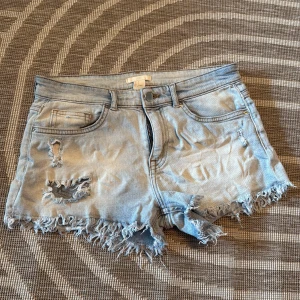 Ljusblå jeansshorts från H&M - Snygga ljusblå jeansshorts från H&M i storlek 36. De har fransiga kanter och slitna detaljer framtill för en cool, avslappnad look. Klassisk femficksmodell med dragkedja och knapp. Perfekta för varma dagar och festivaler.