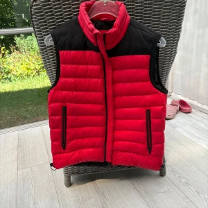 Hugo Boss väst röd/svart, strl S - Säljer en snygg väst från Hugo Boss i storlek S. Röd med svarta detaljer, dragkedjor på fickorna och hög krage. Perfekt som ytterplagg under vår/höst eller som lager under jackan på kyliga dagar. Västen är sparsamt använd och i mycket fint skick – se bild.  Nypris 3200 kr, säljes nu för 450 kr!  Pris: 450 kr Storlek: S Märke: Hugo Boss  Hör av dig vid frågor!