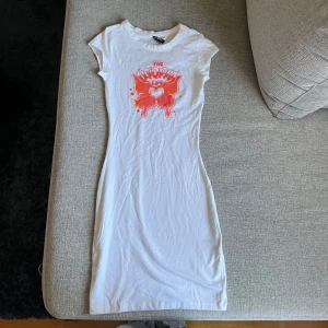 Vit kortklänning från H&M Divided XS - Vit figurnära kortklänning från H&M Divided i storlek XS. Klänningen har korta ärmar och ett tryck framtill i rött och rosa med text och hjärtdetalj. Tillverkad i mjuk bomull och är ungefär till knäna beroende på hur lång man är. Aldrig använd endast testat på.