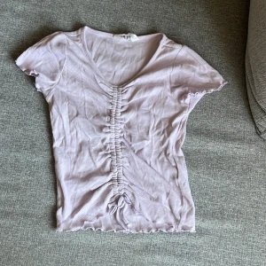 Ljuslila ribbad topp med sydd snörning H&M - Supersöt ljuslila ribbad topp från H&M med ojusterbar snörning. Toppen har v-ringning, korta ärmar och en rosett som detalj. Volangkant nertill och vid ärmslut ger en extra gullig touch. Perfekt för dig som gillar ljusare färger! Mycket mer färgglad i verkligheten än på bild.