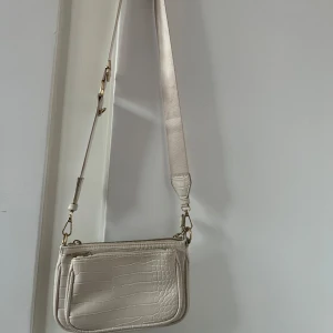 Beige axelväska  - Snygg beige axelväska med krokopräglat mönster och guldfärgade detaljer. Väskan har både bred justerbar axelrem och en kortare kedja för olika stilar. Praktiskt ytterfack och dragkedja upptill. Den har tecken på att den är använd, till exempel några fläckar på insidan 