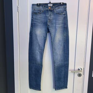 Blå jeans Jack & Jones Comfort Mike - Snygga blå jeans från Jack & Jones i modellen Comfort Mike. Jeansen har klassisk femficksdesign, raka ben och ljusa slitningar framtill för en avslappnad look. Tillverkade i mjuk bomull med orangea kontrastsömmar.