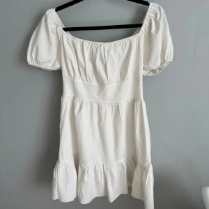 Vit kort klänning med puffärm - Supersöt vit kortklänning med puffiga kortärmar och volang nertill. Klänningen har en offshoulder-modell och är sydd i ett lätt, krinklat bomullstyg som känns luftigt och somrigt. Perfekt för dig som gillar romantisk och trendig stil.