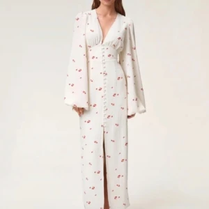 Vit långklänning med blommor från Adoore - Superfin långklänning från Adoore i vit med små röda blommor. Klänningen har v-ringning, långa vida ärmar och knappar framtill. Sitter snyggt i midjan och har en lätt slits framtill. Perfekt för sommarens alla tillfällen!