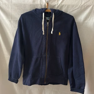 Marinblå hoodie från Polo Ralph Lauren - Snygg marinblå hoodie från Polo Ralph Lauren med dragkedja, huva med vita snören och klassisk gul broderad logga på bröstet. Mjuk bomullskänsla och två stora fickor framtill. Perfekt för chill dagar och streetstyle.