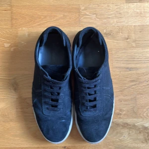 Svarta sneakers från Axel Arigato i mocka - Snygga svarta sneakers från Axel Arigato i mjuk mocka med klassisk låg profil och vita gummisulor. Skorna har rund tå, snörning och diskret logga i guld på sidan. Perfekta för en stilren och clean look. 