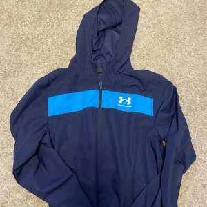 Blå vindjacka från Under Armour - Snygg marinblå vindjacka från Under Armour med huva och dragkedja. Jackan har en blå panel över bröstet med logga och meshfoder på insidan. Perfekt för dig som gillar sportig stil och vill ha något lätt och funktionellt. Jackan är till de med storlek s men passar också xs