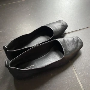 Svarta ballerinaskor från Vagabond - Stilrena svarta ballerinaskor från Vagabond i mjukt skinn. Modellen har fyrkantig tå och platt sula, vilket ger en modern och minimalistisk look. Perfekta för dig som gillar klassiska och tidlösa skor som passar till alla tillfällen!
