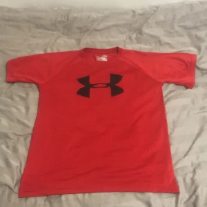 Röd Under Armour t-shirt HeatGear - Röd t-shirt från Under Armour med stort svart logotryck på bröstet. Modellen är loose fit och tillverkad i funktionsmaterial HeatGear som andas, perfekt för träning. Klassisk rund hals och korta ärmar. Enkel och sportig stil.