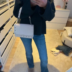 Beige väska med nitar - Snygg beige axelväska i skinn med silverfärgade nitar runt kanterna och kedjedetalj på axelremmen. Från bershka. Stilren och fyrkantig modell som ger en cool vibe till outfiten. Perfekt storlek för det viktigaste och lätt att matcha till olika looks.