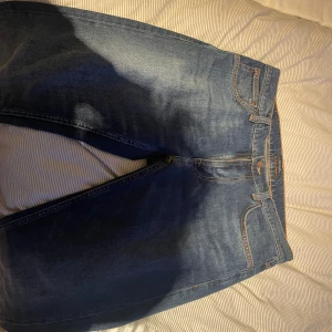 Blå jeans från Nudie Jeans - Snygga blå jeans från Nudie Jeans med klassiska orange sömmar och broderade bakfickor. Jeansen har fem fickor, normal passform och är tillverkade i slitstarkt bomullsjeans. Perfekta för dig som gillar en tidlös och clean look.