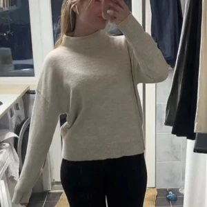 Beige stickad tröja med hög hals - Mysig beige stickad tröja med hög hals och långa ärmar. Tröjan har en avslappnad passform och ribbade muddar vid ärmslut och nederkant. Perfekt för lager-på-lager och enkel att matcha med olika outfits.