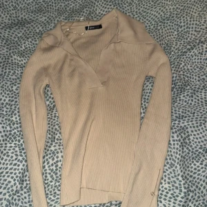 Beige ribbad tröja från Gina Tricot - Säljer en beige ribbad långärmad tröja från Gina Tricot i storlek XS. Tröjan har en snygg krage med v-ringad öppning och är figurnära i modellen. Perfekt att styla med jeans eller kjol för en clean look.