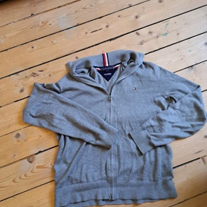 Grå half zip tröja Tommy Hilfiger - Snygg grå tröja från Tommy Hilfiger med half zip och diskret logga på bröstet. Tillverkad i en mjuk blandning av pima bomull och kashmir. Ribbad krage med röd, vit och marinblå detalj. Perfekt för lager-på-lager och chill stil.