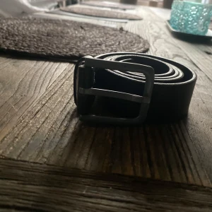 Svart klassiskt bälte i skinn - Stilrent svart bälte i äkta skinn med enkel, matt silverfärgad metallspänne. Bältet har en rak och klassisk form utan mönster, perfekt för att ge en clean look till jeans eller byxor. Passar dig som gillar minimalistisk och tidlös stil.