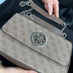Beige axelväska från Guess - Säljer en beige axelväska från Guess med kedjerem och GG-logga i silver framtill. Väskan har ett diskret mönster med logotyptryck över hela ytan och praktiskt fack på baksidan. Perfekt storlek för det viktigaste och snygg detalj till din outfit.