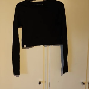 Svart croppad långärmad tröja Röhnisch - Svart croppad långärmad tröja från Röhnisch. Tröjan är ribbad och har en enkel, stilren design med rund halsringning. Perfekt för dig som gillar sportig och clean stil. Materialet är mjukt och stretchigt för extra komfort.