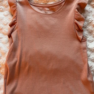 Ribbad volangtopp från H&M, brun - Supersöt ribbad topp från H&M i en varm brun rosa nyans. Toppen är ärmlös med volangdetaljer på axlarna som ger en extra touch. Perfekt för dig som gillar en enkel men ändå trendig stil. Materialet är mjukt och stretchigt för skön passform.