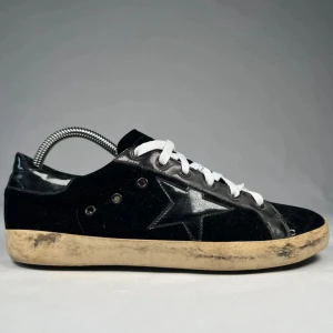 Svarta golden goose skor - Svarta sneakers med vit sula och vita snören. Skorna har en tydlig svart stjärna på sidan och kombinerar mocka och skinn för en cool kontrast. Sulan är beige och har en sliten look som ger en avslappnad streetstyle-vibe. Skorna är det ända som ingår i köpet.