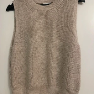 Beige stickad väst  - En stilren beige stickad väst med bred ribbad mudd nedtill och vid halsringningen. Västen är ärmlös och har en rak passform som är lätt att matcha med skjorta eller t-shirt.