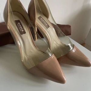 Beige pumps från Bianca x Nelly - Snygga beige pumps med spetsig tå och hög klack från Bianca x Nelly. Skorna har transparenta sidor i syntetmaterial och lackad finish framtill och baktill. Perfekta för dig som vill sticka ut med en modern och trendig look.