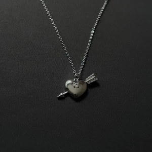 Flèche d’Argent - Silver Hjärthalsband med Pil - Silverpläterat halsband i stainless steel med hjärta och pil, dekorerat med glittrande stenar. Justerbar längd 44 + 4 cm. En tidlös symbol för kärlek med modern elegans