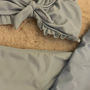 Ljusblå volangbikini från SHEIN - Supersöt ljusblå bikini och kjol från SHEIN med volangdetaljer och knyt framtill på överdelen. Bikinin är i mjukt syntetmaterial och har en clean look utan mönster. Knappast använd och säljer eftersom att den är för liten!!
