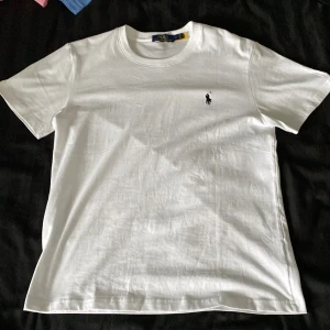 Vit t-shirt från Polo Ralph Lauren - Klassisk vit t-shirt från Polo Ralph Lauren med broderad svart logga på bröstet. T-shirten har rund hals, korta ärmar och är gjord i mjuk bomull. Perfekt basic-plagg med snygg passform och stilren look.