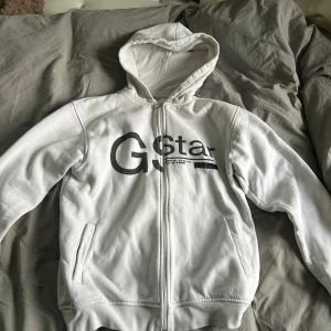 Vit hoodie från G-Star Raw - Vit hoodie från G-Star Raw med stor svart logga framtill och tryck på ryggen. Tröjan har dragkedja hela vägen, huva och två fickor på sidorna. Oversized passform och mjukt material som känns riktigt skönt. Storlek S 