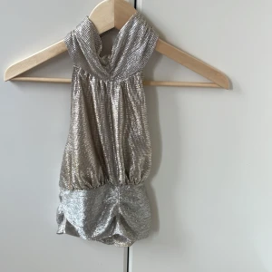Glittrig silver halterneck tubtopp - Säljer en glittrig silverfärgad tubtopp med halterneck och snyggt rynkad nederkant. Toppen har metallic-look och är perfekt för att sticka ut. Sitter tight och har en cool partyvibe.