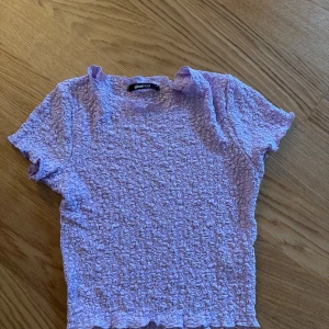 Ljuslila smockad topp från Gina Tricot - Supersöt ljuslila topp från Gina Tricot med smockad struktur och korta ärmar. Toppen har en tight passform och är gjord i ett stretchigt syntetmaterial. Perfekt att styla med jeans eller kjol för en trendig look.