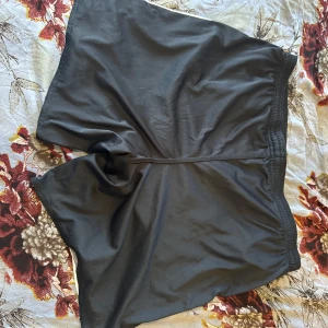 Svarta Umbro shorts med snörning - Svarta sportiga shorts från Umbro med vit kant längs sidorna och klassisk logga på ena benet. De har elastisk midja med snörning för skön passform och är tillverkade i ett lätt och svalt polyestermaterial. Perfekta för träning eller chill dagar.