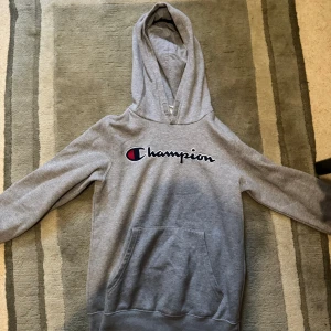 Grå hoodie från Champion - Säljer en grå hoodie från Champion med stor logga broderad på bröstet. Tröjan har huva, magficka och långa ärmar. Perfekt för chill dagar eller när du vill ha en sportig look. Klassisk och enkel design som funkar till det mesta.