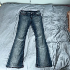 True Religion bootcut jeans blå, stl 28 - Säljer ett par blå bootcut jeans från True Religion med röda kontrastsömmar och klassiska bakfickor med broderi. Jeansen har låg midja och är tillverkade i denim med snygg tvättad effekt. Perfekta för dig som gillar en avslappnad men trendig look.