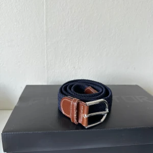 Mörkblå flätat bälte med skinn - Snyggt mörkblått flätat bälte med detaljer i brunt skinn och silvrigt spänne. Bältet har en stilren look med klassisk design och passar perfekt till jeans eller chinos. Flätningen ger en cool textur och skinnet ger en schysst kontrast.