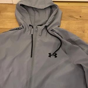 Grå vindjacka från Under Armour - Snygg grå vindjacka från Under Armour med svart logga på bröstet och huva med dragsko. Jackan har hel dragkedja, ventilerande meshpaneler och detaljer med logotyp på axlarna. Perfekt för aktiva dagar och sportiga outfits.