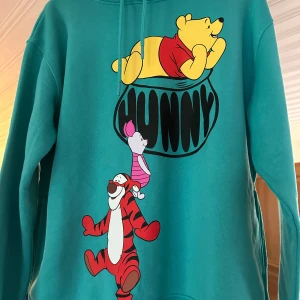 Turkos Disney hoodie med Nalle Puh - Turkos hoodie från Disney med tryck av Nalle Puh, Nasse och Tiger på framsidan. Tröjan har huva med snörning och är långärmad. Perfekt för dig som älskar klassiska Disney-karaktärer och vill ha en färgglad och bekväm stil.