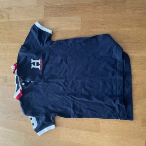 Marinblå Hilfiger pike med logga - Snygg marinblå pike från Tommy Hilfiger med stor H-logga och broderad text på bröstet. Piken har korta ärmar med vita och röda detaljer på kragen och ärmsluten. Perfekt för en sportig och stilren look.