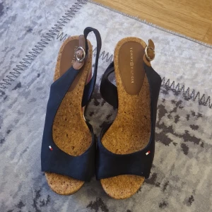Tommy Hilfiger kilklack sandaler, strl 39 - Snygga sandaler från Tommy Hilfiger med kilklack i kork och ovandel i mörkblå bomull. Sulan har ränder i rött, vitt och blått. Öppen tå och justerbar rem med spänne. Perfekta för sommaren och ger en fräsch look till din outfit. Använda fåtal gånger som nya