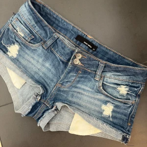 Lågmidjade jeans shorts - Snygga blå jeansshorts med råa kanter och slitna detaljer framtill och baktill. Klassisk femficksmodell med låg midja och knappgylf. Perfekta för varma dagar och en avslappnad stil.