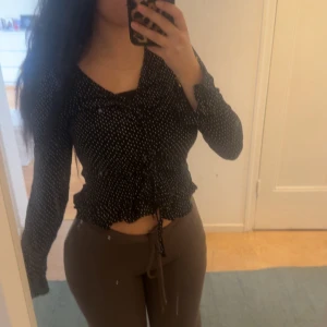 Svart blus med vita prickar från H&M - Säljer en svart blus från H&M med vita prickar. Blusen har lång ärm, v-ringning och knytning i midjan. Den är croppad och har en lätt volangkant nertill. 