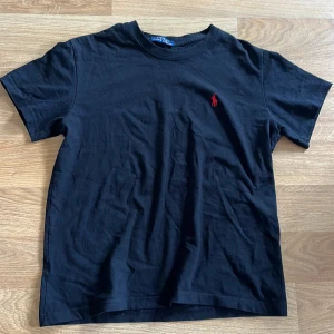 Svart t-shirt från Ralph Lauren - Svart t-shirt från Ralph Lauren med klassisk passform och röd broderad logga på bröstet. Perfekt basic-plagg med rund hals och korta ärmar. Stilren och enkel design som funkar till det mesta.