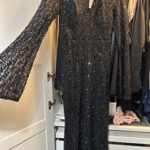 Svart paljettklänning från Vera & Lucy - Säljer en lång svart klänning från Vera & Lucy med v-ringning och långa ärmar. Klänningen är täckt av svarta paljetter som ger en glittrig look och har en figurnära passform. Perfekt för dig som vill sticka ut på festen!