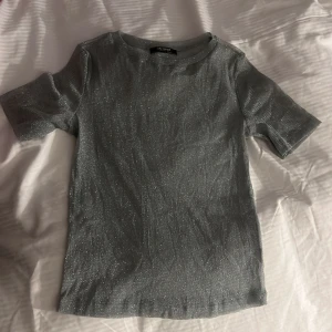 Genomskinlig festtopp - Säljer en glittrig silverfärgad topp från Zara Trafaluc. Toppen har korta ärmar och är tillverkad i ett tunt, transparent material som ger en snygg glansig effekt. Perfekt för att sticka ut med en trendig look.
