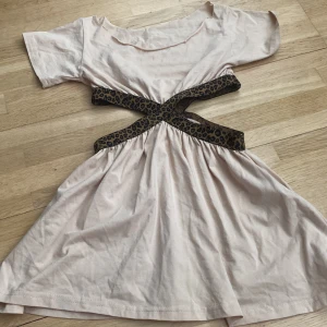 beige lång blus med leoparddetaljer - Säljer en beige blus med korta ärmar och unika korsade leopardmönstrade band i midjan. blusen har en rund urklippt halsringning och ett utställt kjolparti. Perfekt för dig som vill sticka ut med coola detaljer!