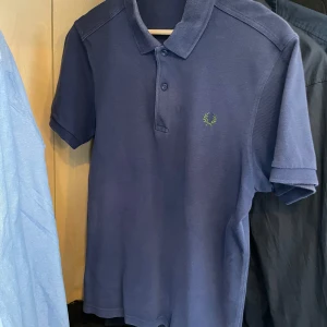 Blå piké t-shirt från Fred Perry - Säljer en klassisk blå piké t-shirt från Fred Perry med krage och knappar framtill. T-shirten har korta ärmar och den ikoniska Fred Perry-loggan broderad i grönt på bröstet. Perfekt för en stilren och avslappnad look.