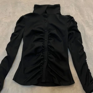 Svart långärmad skjorta med rynkade detaljer - Snygg svart skjorta med hög krage och långa ärmar. Skjortan har knappar framtill och rynkade detaljer längs knappraden som ger en cool och trendig look. Perfekt för dig som gillar stilrena plagg med en twist.