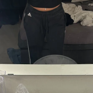 Svarta mjukisbyxor från Adidas - Svarta mjukisbyxor från Adidas med vit logga på ena benet och dragsko i midjan. Byxorna har en avslappnad passform och är perfekta för chill eller träning. Klassisk design med resår i midjan och raka ben.