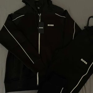 Svart hoodie med dragkedja från BOSS - Säljer en svart hoodie från BOSS med vit dragkedja och vita detaljer längs ärmarna. Tröjan har huva med snörning och BOSS-logga på bröstet. Perfekt för en sportig och stilren look.